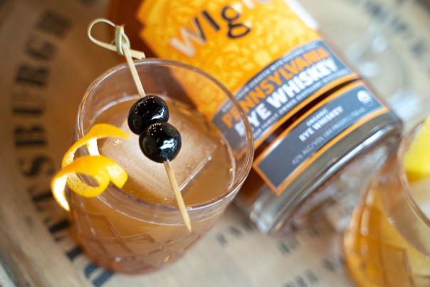 A glass of whiskey with a large ice cube, sliver of orange peel and two berries on a toothpick