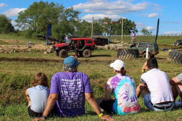 Group of six people sitting on grass watching trucks drive through an obstacle course on a sunny day.