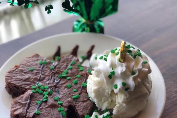 A brownie with green sprinkles on a plate with vanilla ice cream and more green sprinkles and hot fudge drizzle on the brownie