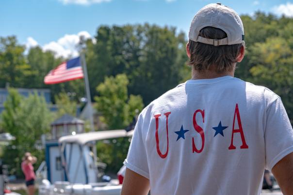 A man wearing a USA shirt looks at an American flag at a lake in the Poconos.