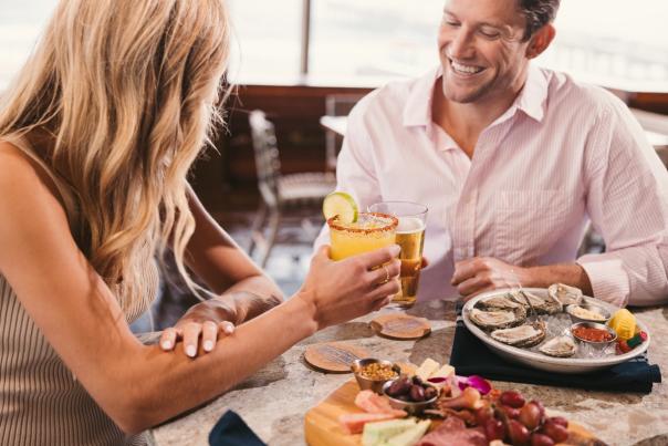 a man and woman toasting with two drinks - their table has a plate of oysters on the half-shell and  a charcuterie board