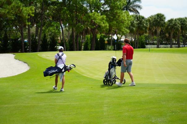 A father and son walking on a beautiful, green golf course in Pompano Beach. Dad's bag is on a push cart, and the son is carrying his bag.