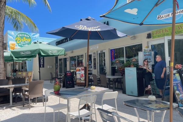 A strip of restaurants with outdoor tables with umbrellas, on a palm-tree-lined street in sunny Pompano Beach.