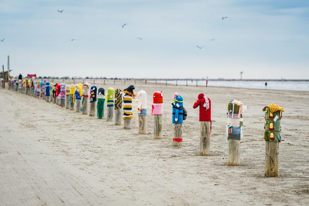 Row of wooden bollards decorated as "Bollard Buddies" with colorful outfits on a sandy beach.