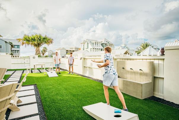 A person tosses a bean bag during a cornhole game on an outdoor turf patio with white fencing.