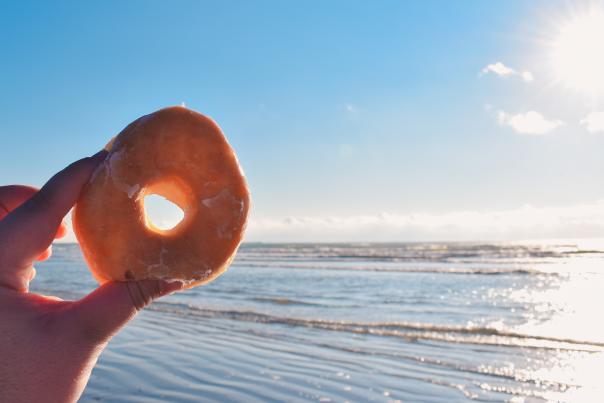 A donut is held up in front of a beach scene.