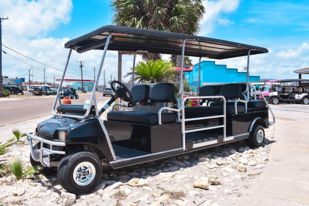 A golf cart sits on rocks. It is long and black and has space in the middle for a wheelchair.