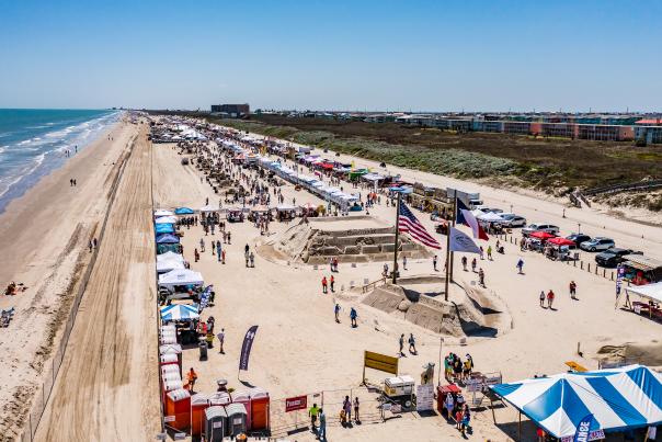 Aerial photo of a festival on the beach featuring sand sculptures, vendors, and large flags.
