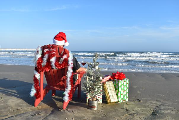 A man in a santa hat sitting in a chair on the beach facing the ocean with Christmas gifts next to him and a tiny Christmas tree.