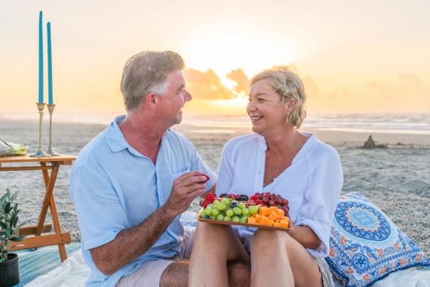 An older man and women sitting on the beach having a picnic with blue blankets and pillows. They are sharing a tray with grapes and cheese while the sun sets behind them.