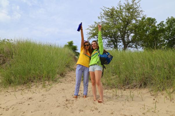 Two young teens hold their arms up in celebration while standing in the sand in front of vegetation.