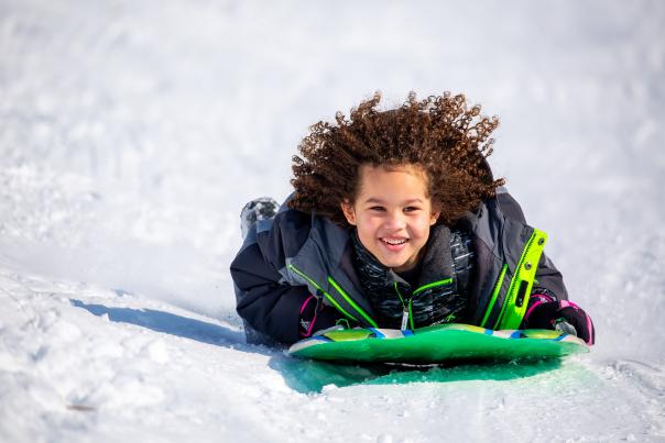 A young, smiling child sleds down a snowy hill on a green sled. Curly hair is swept back on the child's face.