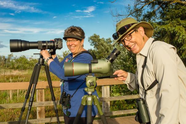 Two women stand in front of the birding scopes on tripods.
