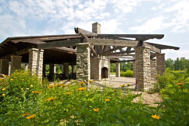 A stone and wooden pavilion with a fireplace sites in the middle of yellow flowers and greenery.