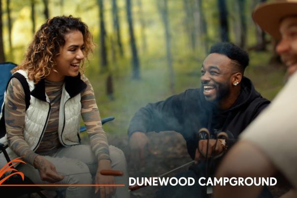 A woman and man look at each other laughing while sitting in front of a campfire.