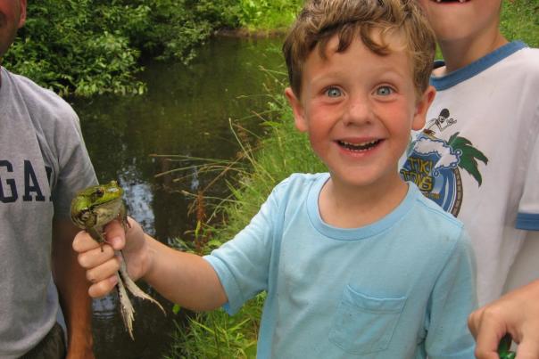 A small child in a light blue shirt holds a frog in his right hand. He is smiling broadly.