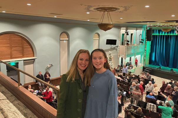 A woman and a young girl stand together, smiling at the camera inside a theater balcony. The woman wears a dark green coat, while the girl wears a long-sleeved blue dress with a black headband. Behind them, a large audience fills the lower-level seats, and the theater's ornate decor, including chandeliers, balcony railings, and a stage with blue curtains, is visible. The space is decorated with holiday lights, suggesting a festive event.