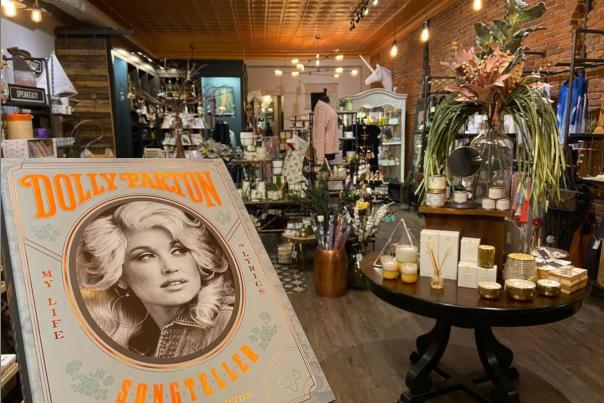 A Dolly Parton book is being held up in front. A boutique store is in the background. Tables with items such as candles and gifts are in the scene.