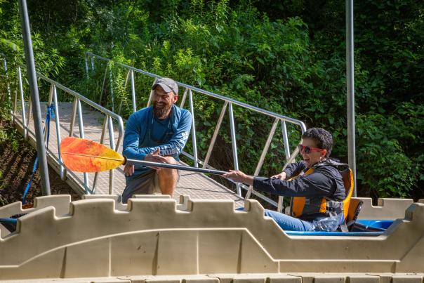 A man kneels down to help a woman in a kayak pulling up onto a launch.