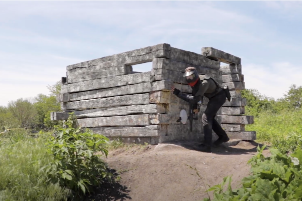 A man dressed for paintball hides behind a rustic wooden structure.