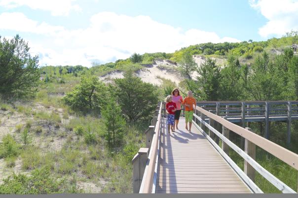 A woman and two small children hike along a wooden boardwalk over vegetation-covered dunes.
