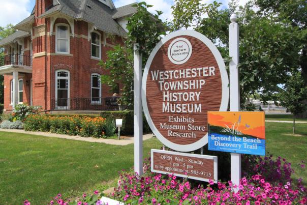 An oval sign reads "Westchester Township History Museum." In the background is a historic brick building. Pink flowers are around the sign.