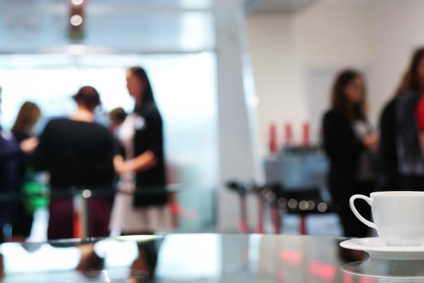 A photograph showing a cup and saucer in the foreground, with people in smart clothes in the background at a conference event