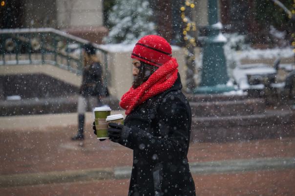 Woman in a red winter hat and scarf walking in the snow holding two cups of coffee.