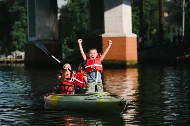 A woman and 2 children kayaking, the child in the front is standing with his arms above his head excited