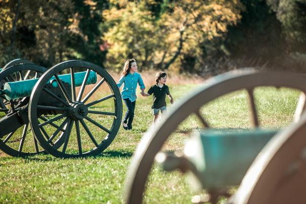 Two kids holding hands, walking on the field next to cannons
