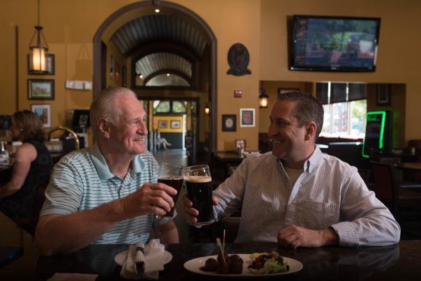 2 men smiling, drinking beer, sitting at a bar with a plate of food on the bar