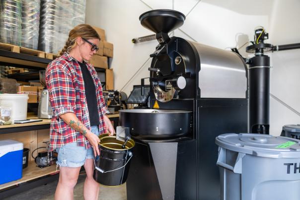 A woman in a casual clothes stands next to a coffee roaster catching beans in a tin