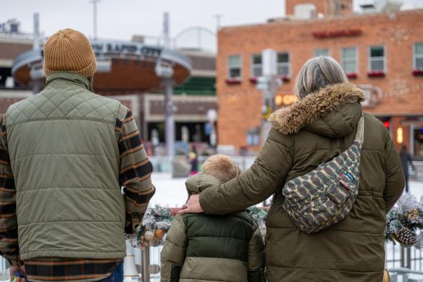 A family wearing winter gear faces away from the camera toward the Ice Rink at Main Street Square. Mom, in a green winter jacket and wearing a patterned backpack, has her arm around her son, wearing an olive green winter jacket in the middle of the group.