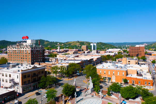 Aerial view of downtown Rapid City featuring historic brick buildings, tree-lined streets, and the Hotel Alex Johnson sign, with green Black Hills in the background under a clear blue sky.