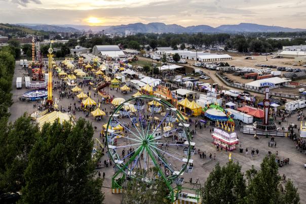 Aerial view of a bustling fairground at sunset, featuring a large Ferris wheel, colorful tents, and various rides. People are scattered throughout the vibrant scene, adding a lively atmosphere.
