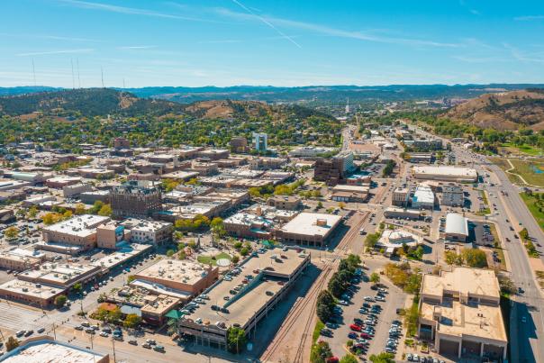 Aerial view of downtown Rapid City with buildings, roads, and parking lots. Surrounding green hills and a clear blue sky create a serene and expansive atmosphere.