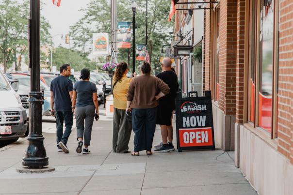 Downtown Rapid City Ambassador in a yellow shirt talks with visitors outside a storefront along a busy sidewalk with parked cars, street banners, and a “Now Open” sign.