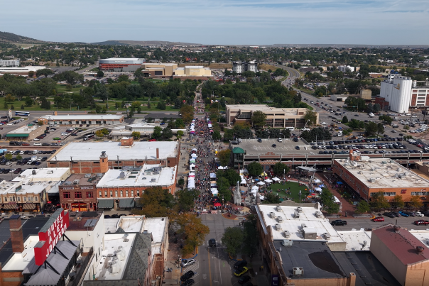 An aerial view of the Pumpkin Festival footprint, looking north from the Hotel Alex Johnson up 6th Street. The street is filled with colorful tent tops and crowds of people throughout.