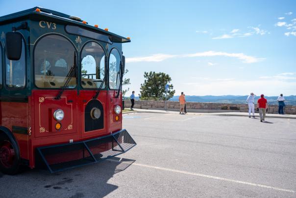 A red-and-green City View Trolley is parked at Stonewall Overlook, where visitors stand along a stone wall overlooking a scenic mountain landscape beneath a bright blue sky.