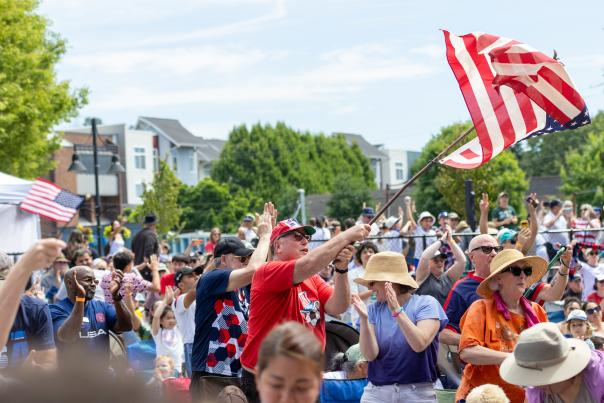 A crowd gathered at an outdoor Independence Day celebration in Renton, waving American flags during a community event with spectators lining the street and local festivities underway.