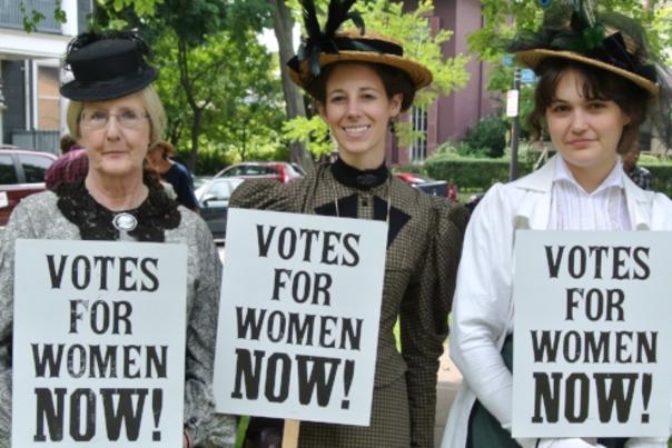 three women hold votes for women now signs