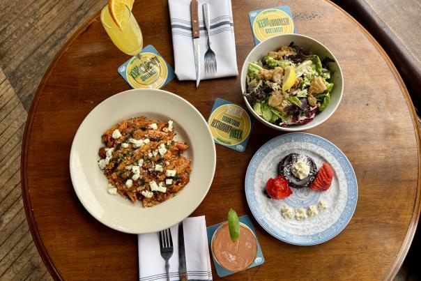 A shot of a full table from above. There is a bowl of pasta, a salad, a brownie on a decorated plate, and an orange and pink drink in two tall cocktail glasses.