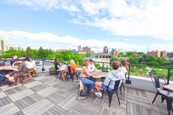 Photo of outdoor rooftop dining at the Genesee Brew House with blue skies and clouds, High Falls waterfall, buildings in the Rochester city skyline, and people sitting at tables on the patio