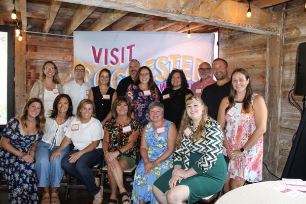 Group photo of smiling men and women in front of colorful backdrop that says "Visit Rochester"