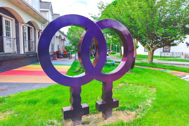 A large purple sculpture of intertwined female symbols stands on green grass in front of Rochester's Forum 73, with a rainbow sidewalk and trees in the background.