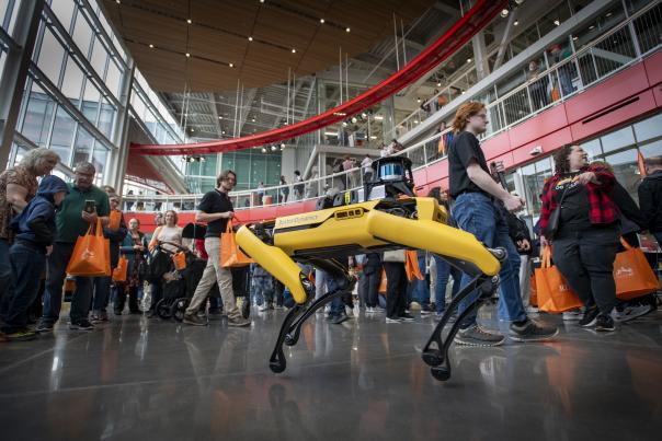 An image of a four-legged robot with a yellow "body" and black "legs" surrounded by people visiting the Imagine RIT festival