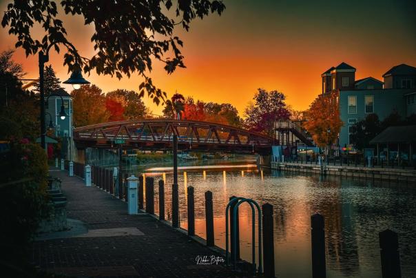 A shot of the Erie Canal in Fairport, NY. The Main Street Bridge can be seen backlit by the orange glow of a setting sun. Trees featuring fall foliage can be seen in the foreground and background of the photo.