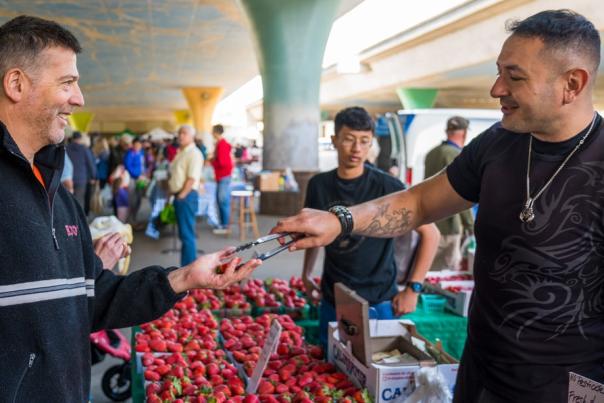 A group of people at a market, one man handing another man a strawberry to sample