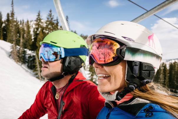 A man and a woman wearing helmets and goggles riding the ski lift at Salt Lake's ski resorts