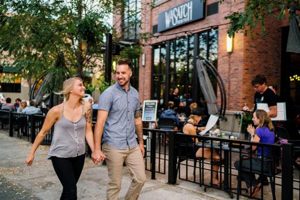 Man and woman holding hands walking past some diners on a patio in the Sugarhouse neighborhood